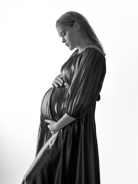 Μια έγκυος κοπέλα με ξανθά μαλλιά σε ένα μακρύ φόρεμα θέτει μπροστά από την κάμερα σε ένα ελαφρύ φόντο. Η έννοια της ευτυχισμένης εγκυμοσύνης. Ασπρόμαυρη φωτογραφία. - Φωτογραφία, εικόνα