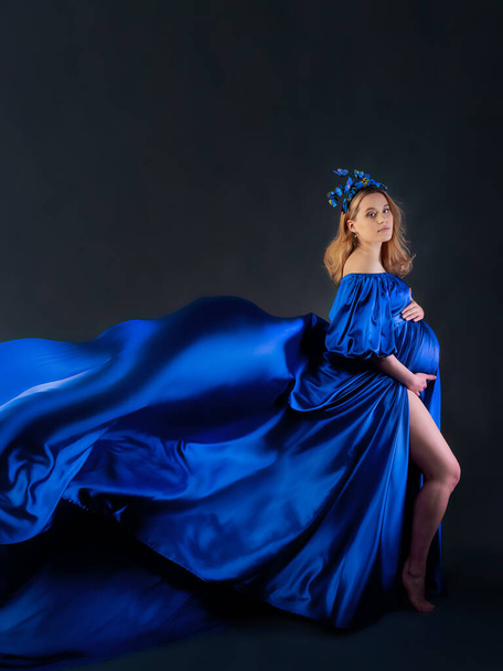 Вагітна дівчина з світлим волоссям у довгій блакитній сукні з протікаючим потягом і пов'язкою з позуваннями метеликів перед камерою. Концепція щасливої вагітності
 - Фото, зображення