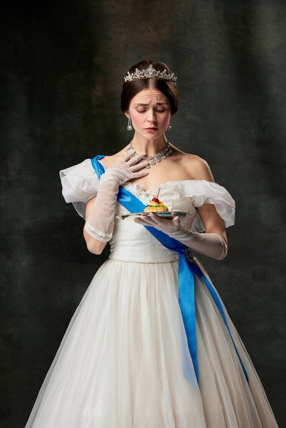 Royal diet. Elegant sad woman, royal person, queen or princess in white medieval outfit cant tasting cake on dark vintage background. Concept of comparison of eras, modern sweets, fashion, beauty, ad - Foto, Imagem
