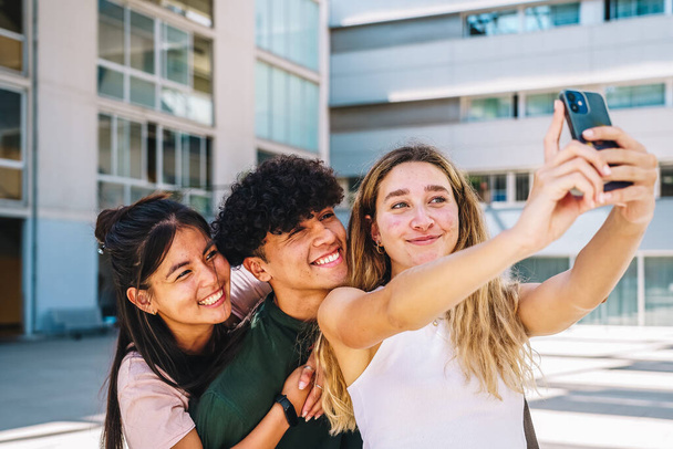 Iloinen nuori nainen, jolla on pitkät vaaleat hiukset hymyilevät samalla kun selfie älypuhelimella iloinen latino mies- ja naispuoliset ystävät seisovat kampuksella aurinkoisena päivänä. - Valokuva, kuva