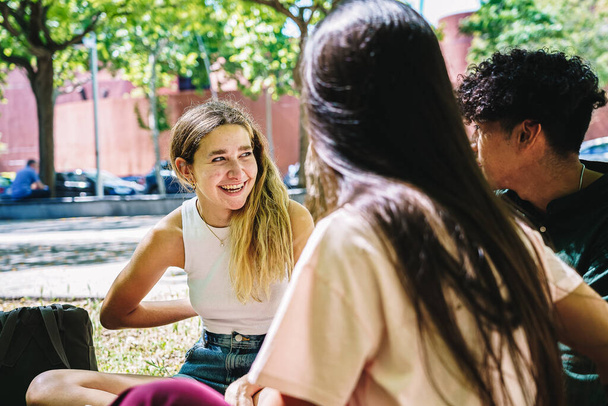 Ευτυχισμένος νεαρός πολυεθνική άνδρες και γυναίκες καλύτεροι φίλοι σε casual ρούχα χαμογελώντας και μιλώντας, ενώ κάθεται στο γρασίδι στην πανεπιστημιούπολη την ηλιόλουστη μέρα - Φωτογραφία, εικόνα