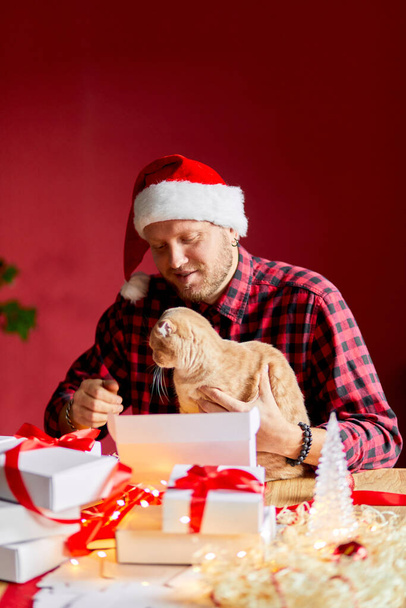 Vet Man in Santa hat and cat in hand making greeting card for New Year and Christmas, congratulation for clients or friends or family , packing presents or gifts, mail manager concept. - Foto, afbeelding