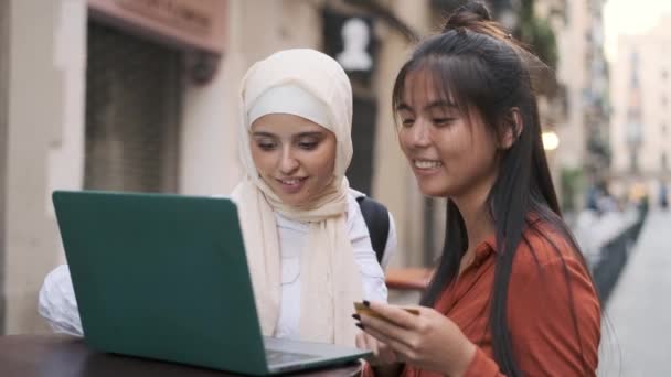 Die multiethnischen jungen Frauen benutzen eine Kreditkarte, während sie online mit einem Laptop auf der Straße einkaufen. Technologiekonzept. - Filmmaterial, Video