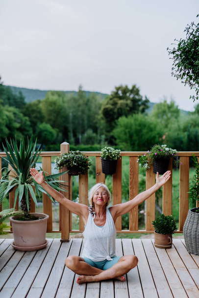 Senior woman sitting outdoors on a terrace in summer, doing yoga exercise. - Foto, afbeelding