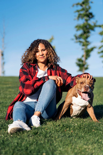 Cheerful african woman with curly hair sitting in the park next to her adorable dog and looking at camera. - Zdjęcie, obraz