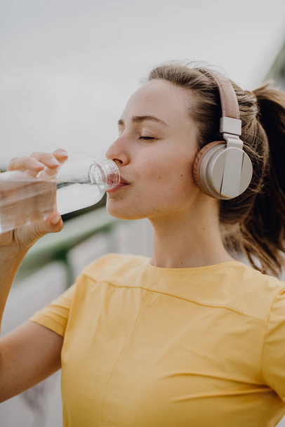 Young woman drinking water and listening the music trough headphones, during jogging in city, healthy lifestyle and sport concept. - Foto, afbeelding