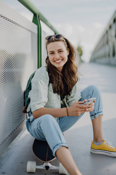 Happy young woman sitting on a skateboard at city bridge. Youth culture and commuting concept. - Foto, Imagem