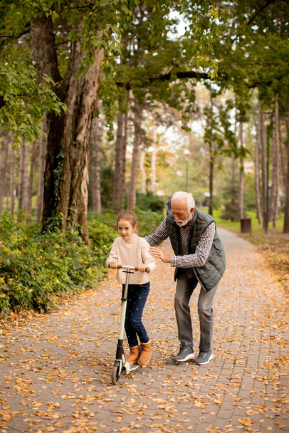 Hombre mayor enseñando a su nieta a montar patinete scooter en el parque de otoño - Foto, imagen