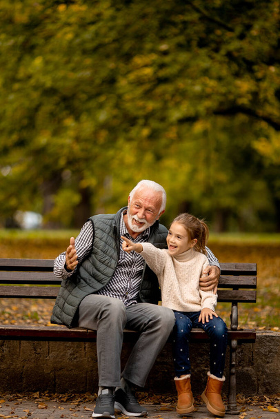 Όμορφος παππούς περνάει χρόνο με την εγγονή του στο παγκάκι την ημέρα του φθινοπώρου. - Φωτογραφία, εικόνα