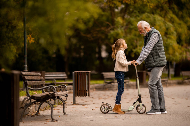 老人は孫に秋の公園でキックスクーターに乗る方法を教えています - 写真・画像