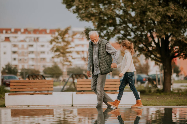 Handsome grandfather spending time with his granddaughter by small water pool in park on autumn day - Fotó, kép