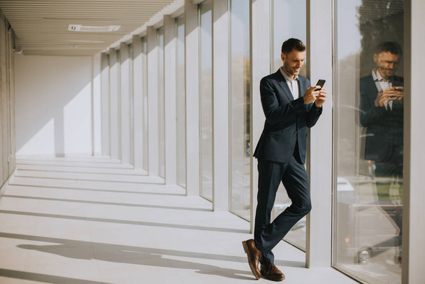 Красивый молодой бизнесмен, использующий мобильный телефон в современном офисном коридоре - Фото, изображение