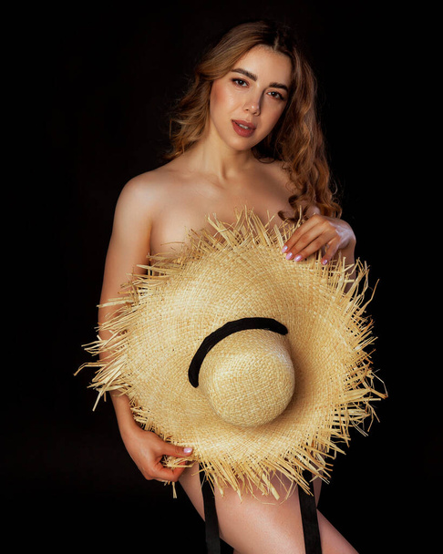 Naked woman cover herself by a big straw hat, on black background in studio - Foto, Imagem