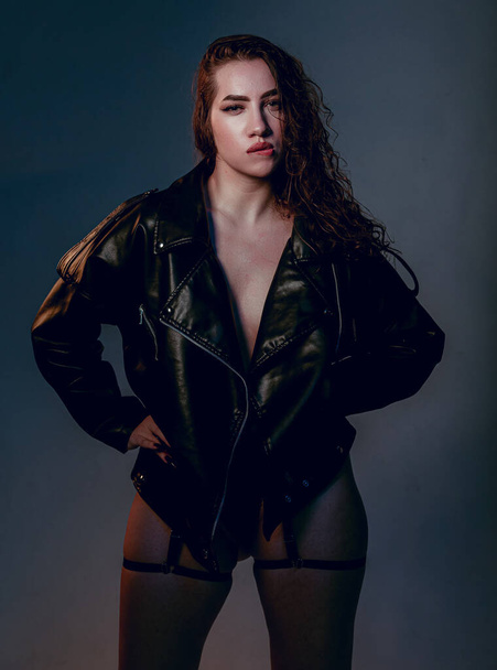 Plus size woman in black leather jacket. Beautiful curvy body, seductive lady posing In underwear. Attractive oversized female In lingerie standing on dark blue studio background. - Valokuva, kuva