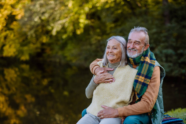 Coppia di anziani innamorati seduti insieme su una panchina vicino al lago, durante la giornata autunnale. - Foto, immagini