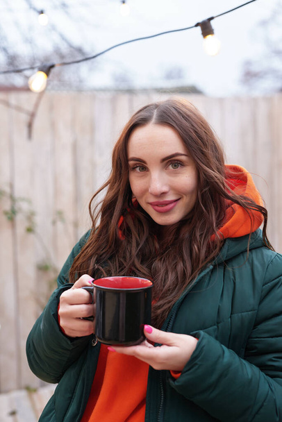 Young woman on background of Christmas tree with cup of coffe outdoor. Backyard decoration new year - Foto, Imagem
