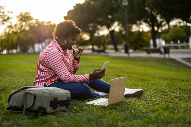 Young afro curvy woman using her phone on a lunch break, eating a toast sandwich on the grass in the park in front of the opened laptop - 写真・画像