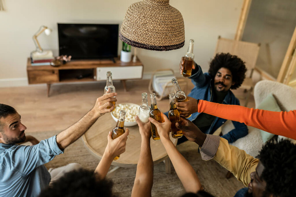 Grupo multirracial de hombres colgando en casa y brindando con botellas de cervezas - Foto, imagen