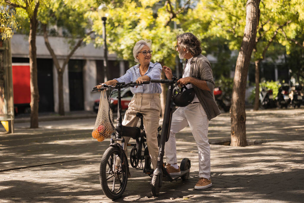 市場から果物を購入した後、素敵な先輩夫婦が会話をしています。女性はメッシュバッグで自転車に乗って男は電動スクーターに乗っています - 写真・画像