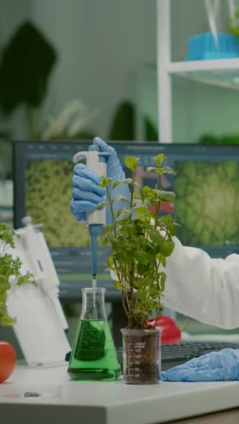 Vídeo vertical: Científico biólogo hablando de la solución de frasco médico que pone en el plantón verde para el experimento genético. mujer investigadora en bata blanca que trabaja en laboratorio de microbiología profesional - Imágenes, Vídeo