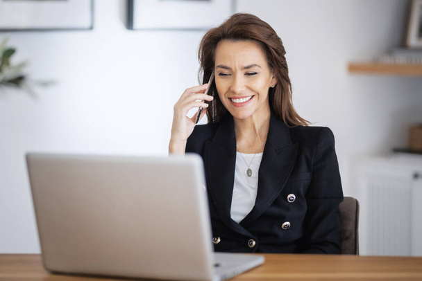 Shot of an attractive businesswoman using a laptop and having a call while working in her home office. Confident female wearing blazer.  - Foto, Imagem