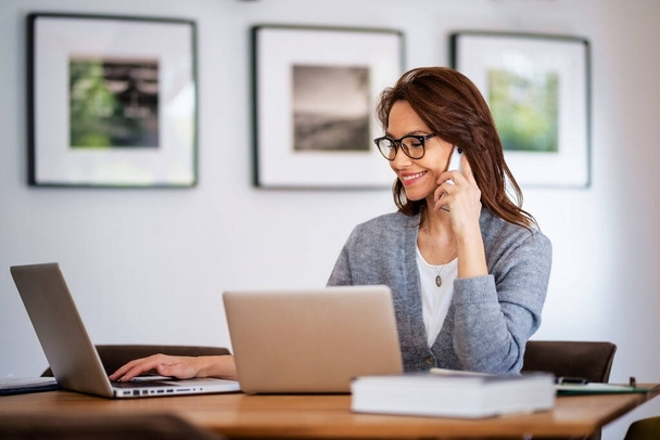 Attractive woman wearing eyewear and casual clothes while working from home. Business woman using laptops and making a call. Home office. - Photo, image
