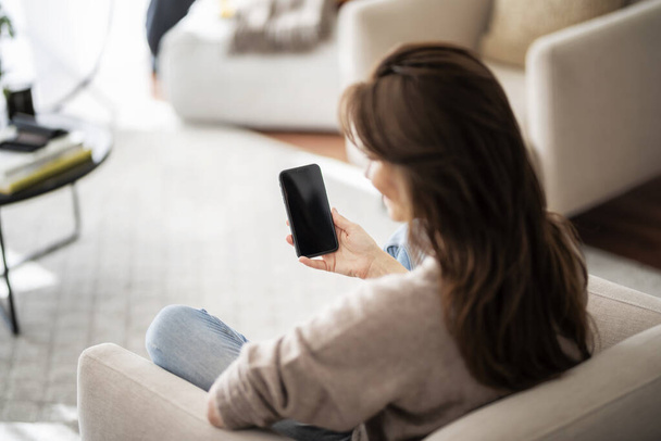 Rear view shot of an unrecognizable woman holding smartphone in her hand while relaxing at the armchair. Blank screen template mock up. Over the shoulder view. - Foto, immagini