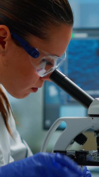 垂直ビデオ:医学開発研究所で顕微鏡下を見ている科学者の肖像,ペトリ皿のサンプルを分析.最先端の医薬品研究所でウイルスを調べるバイオテクノロジー研究 - 映像、動画