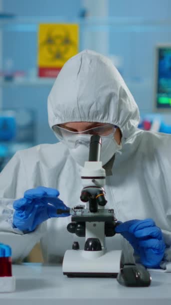 Vídeo vertical: Científico en traje ppe haciendo ajustes y mirando a través del microscopio de laboratorio, escribiendo en pc. Químico en mono que trabaja con varias bacterias, muestras de sangre de tejido para - Metraje, vídeo