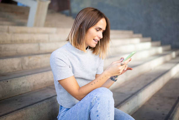 Seitenporträt einer Frau, die auf der Treppe sitzt und auf ihr Handy schaut  - Foto, Bild