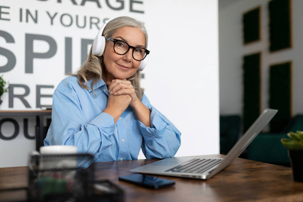 exitosa mujer gerente viendo video de entrenamiento en el ordenador portátil con auriculares sentados en la mesa en la oficina. - Foto, Imagen