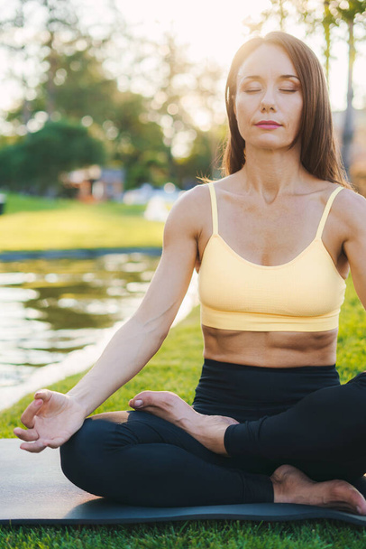 Brunetka kobieta w odzieży sportowej siedzi w pozycji lotosu z zamkniętymi oczami ciesząc się medytacją w słoneczny poranek w przyrodzie. Trening. Zdrowa aktywność - Zdjęcie, obraz