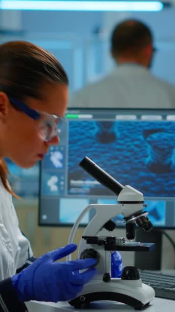 Pionowe wideo: Naukowiec patrzący na biologiczne próbki pod mikroskopem wpisujący na pc. Zespół chemików pracujących w laboratorium, badających ewolucję wirusa przy użyciu zaawansowanych technologii do badań naukowych szczepionki - Materiał filmowy, wideo