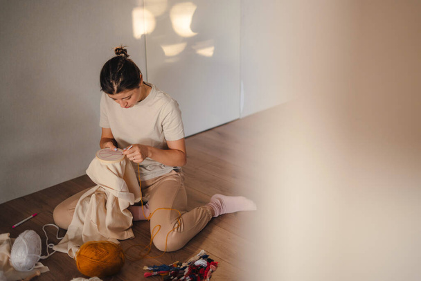 Zaostřená mladá žena šije v pohodlí svého bytu a vyšívá barevný obraz do smyčky. Textilní látky. Domácí dekorace. Design domů. - Fotografie, Obrázek