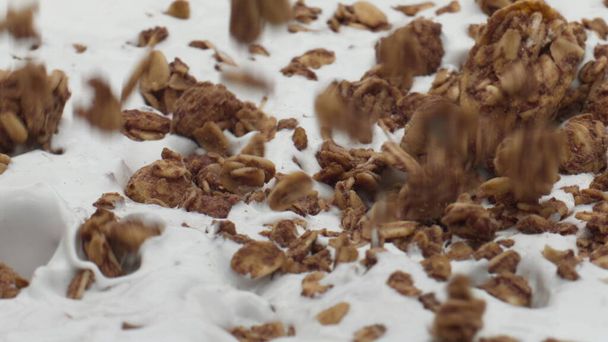Verter la sabrosa granola en yogur natural fresco en cámara súper lenta de cerca. Copos de avena al horno que caen sobre la superficie cremosa del postre. Delicioso ingrediente de trigo para preparar el desayuno nutritivo. - Foto, imagen