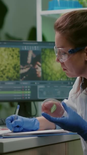 Vídeo vertical: Mujer bióloga escribiendo experiencia médica mientras sostiene la placa de Petri con carne de res vegana en manos que trabajan en el laboratorio de microbiología. Mujer química investigando alimentos modificados genéticamente - Imágenes, Vídeo