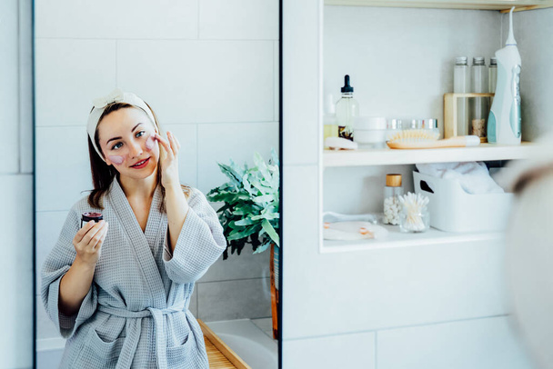 Young woman in bathrobe looking in the mirror and applying facial natural cosmetic clay mask on her face in bathroom. Cosmetic procedures for skin care at home. Beauty self-care. Selective focus. - Фото, изображение