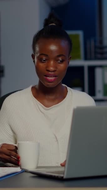 Black business woman using wireless headphone drinking coffee duing video conference working overtime from start-up office in front of laptop. Freelancer using talking on virtual meeting at midnight - Footage, Video