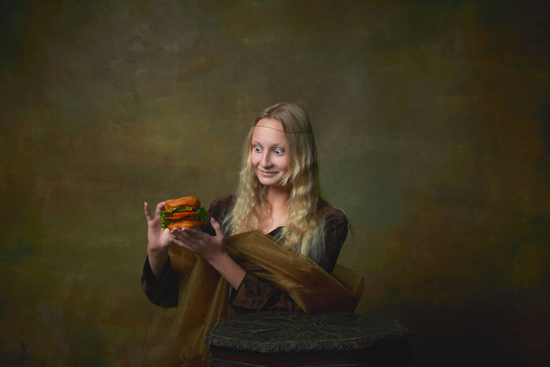 Surprised young girl as Mona Lisa picture tasting burger over dark vintage background. Retro style, art, fashion, comparison of eras concept. Beautiful female model like classic historical character - Фото, изображение