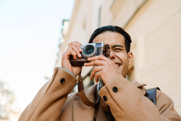 Young man wearing coat taking photo on retro camera at city street outdoors - Valokuva, kuva