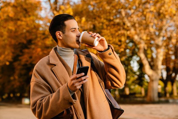 Young man wearing coat using cellphone and drinking coffee while walking in autumn park - Foto, Imagem