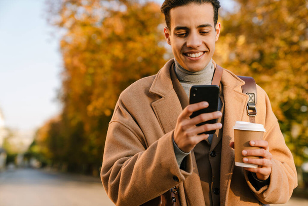 Молодой человек в пальто с помощью мобильного телефона и пить кофе во время прогулки в осеннем парке - Фото, изображение