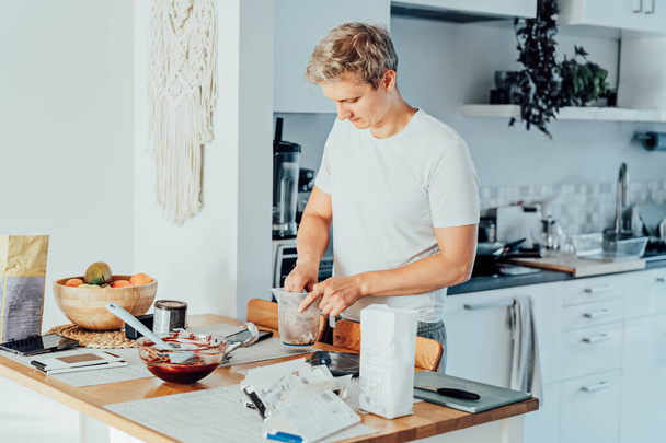 Young man cooking brownie cake on the light modern kitchen at home. Preparing steps for baking cake. Culinary hobby. Cooking online video classes on smartphone. Selective focus. copy space. - Foto, imagen