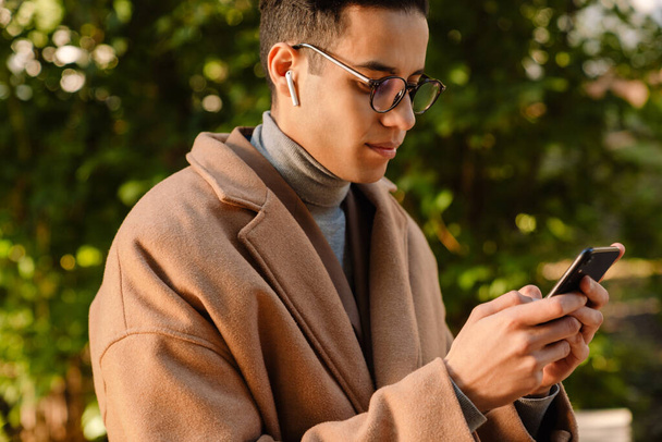 Young man wearing glasses using mobile phone and wireless earphones in park - Photo, Image