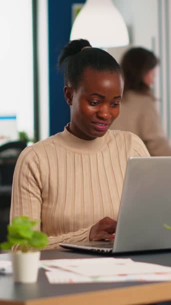 Boldog fekete menedzser olvasás feladatokat laptop és gépelés ül az íróasztalnál elfoglalt start-up iroda, míg a különböző csapat elemzi a statisztikai adatokat a háttérben. Többnemzetiségű csoport a projektről - Felvétel, videó