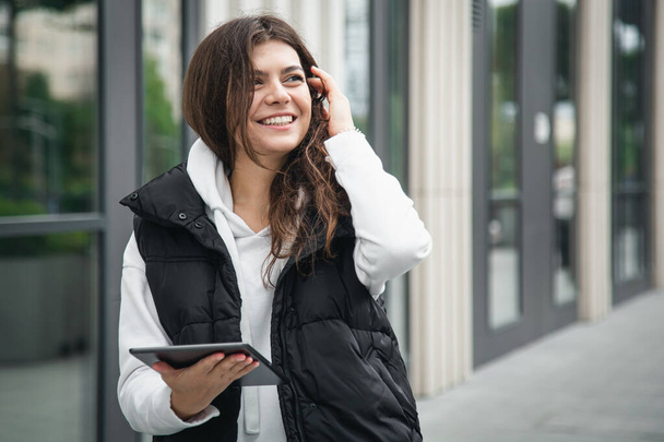 Zakelijke jonge vrouw staat buiten en gebruikt een tablet tegen een wazige achtergrond van een gebouw. - Foto, afbeelding