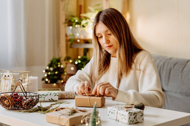 Vrouw wikkelt kerst eco natuurlijke geschenkdozen thuis. Cadeaus bereiden op witte tafel met decor elementen, verlichting en items Kerst of Nieuwjaar DIY verpakking Concept. - Foto, afbeelding