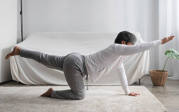 Señora mayor de pie ejercicio haciendo Yoga pose en la sala de estar. actividad de mediana edad para equilibrar el cuerpo - Foto, imagen