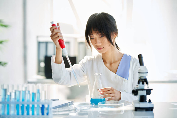 Cientista feminina fazendo pesquisa microbiológica usando pipeta, frasco e tubos de ensaio que trabalham em laboratório químico moderno. Medicina, conceito de biotecnologia - Foto, Imagem