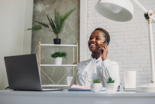 Afro-Amerikaanse uitvoerend manager accountant met behulp van computer tijdens het praten op de telefoon in het kantoor. Communicatie met leveranciers. Millennial zakenvrouw portret. - Foto, afbeelding